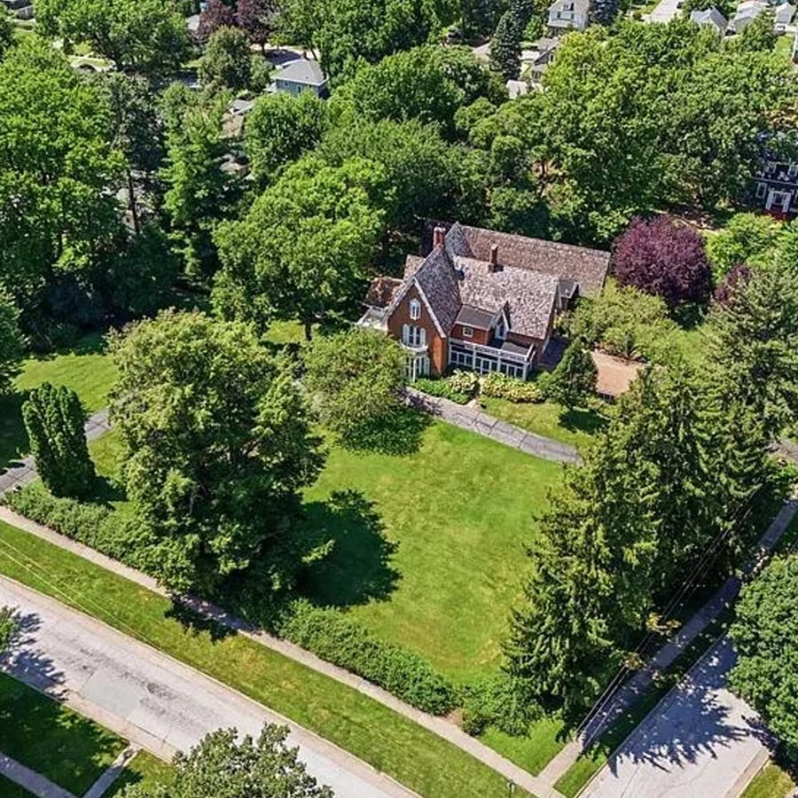 Aerial view of a large brick house surrounded by green trees and spacious lawns, located at a street corner in a suburban neighborhood.