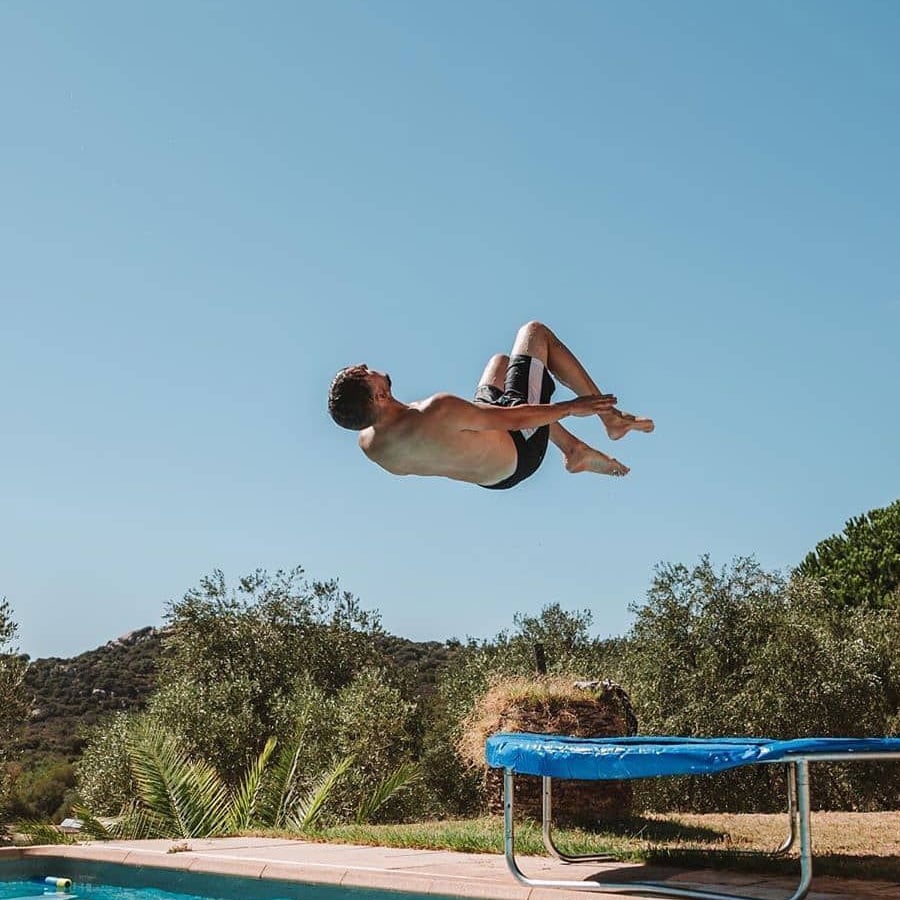 A person in swim trunks is mid-air, performing a backflip above a blue trampoline near a pool, with trees and hills in the background under a clear blue sky.