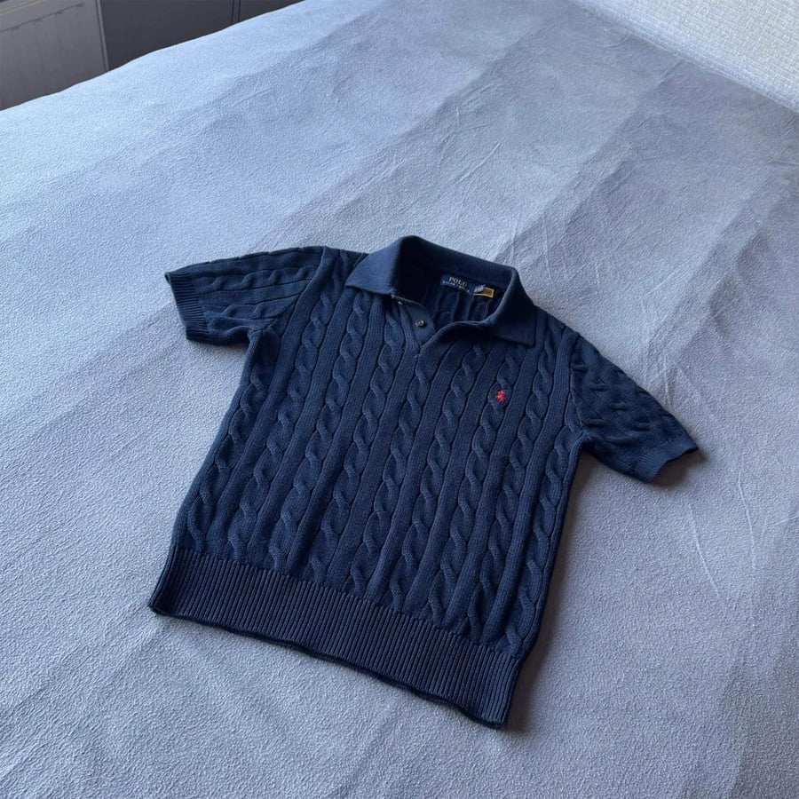 A navy blue short-sleeve cable knit polo shirt with a collar and a small red emblem on the chest, neatly laid out on a gray bedspread.