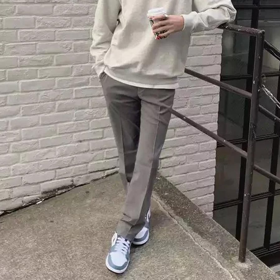 A person in a light sweatshirt and gray pants stands against a white brick wall, holding a coffee cup, with one leg crossed over the other and wearing white sneakers.