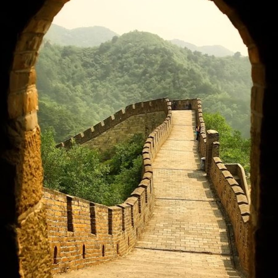 View of the Great Wall of China winding through green hills, seen through a stone archway on a sunny day.