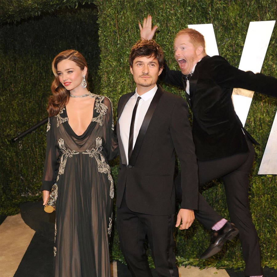 A woman in a long, dark gown and a man in a black suit pose on a red carpet while another man photobombs them by jumping and making a funny face in the background.