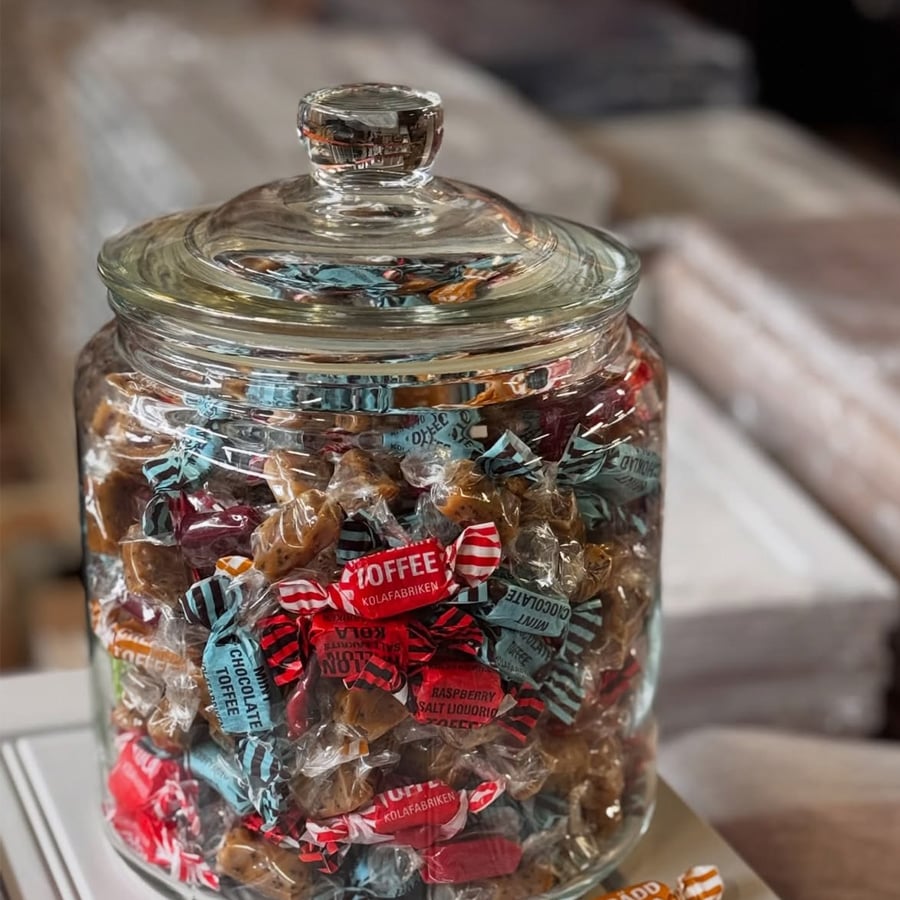 A clear glass jar with a lid filled with assorted wrapped toffees in blue, red, and orange wrappers, sitting on a white surface.