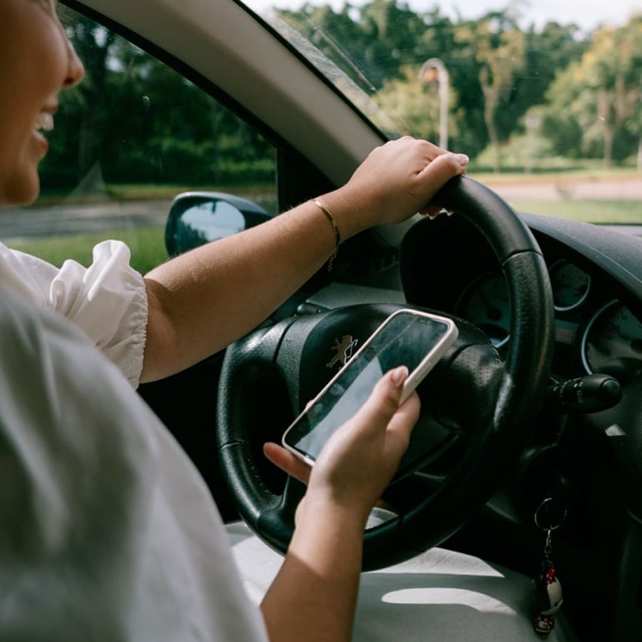 A person driving a car holds the steering wheel with one hand and uses a smartphone with the other, while smiling. Trees and greenery are visible outside the window.