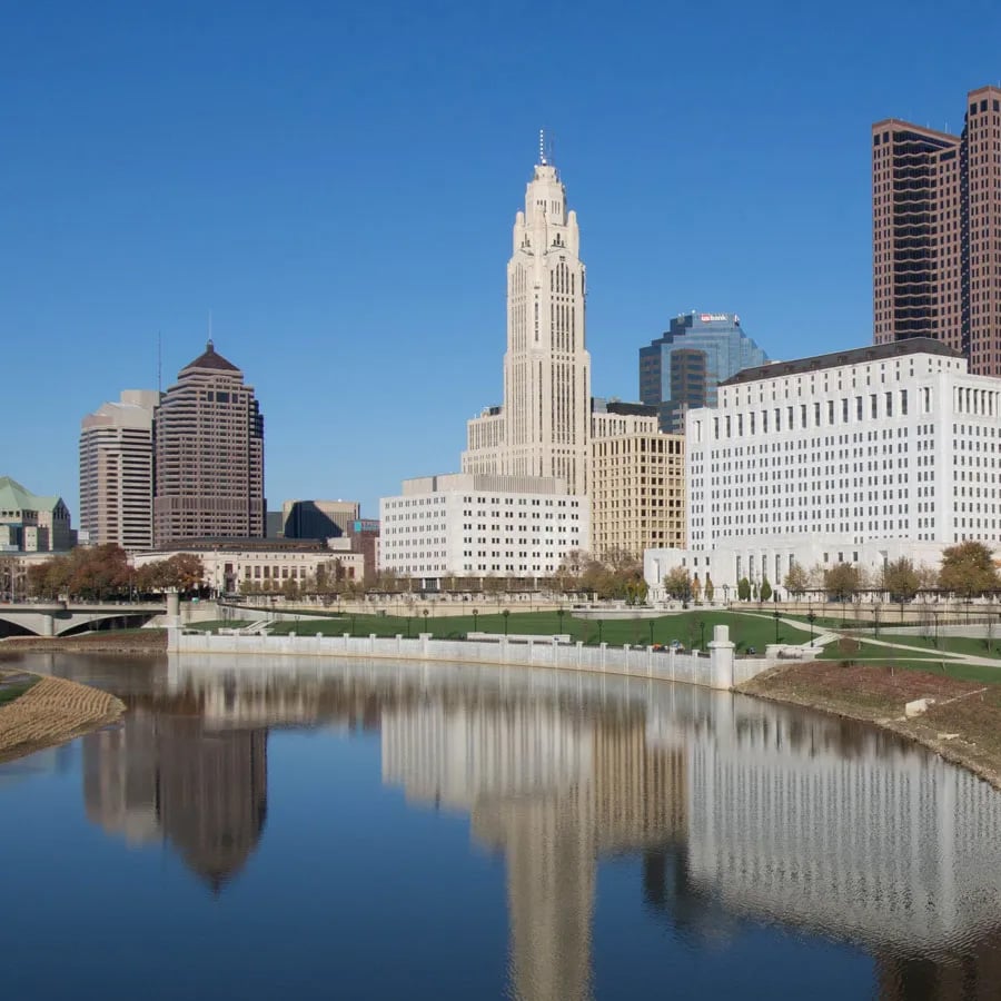 Downtown Columbus, Ohio skyline with tall buildings and clear blue sky reflected in the Scioto River on a sunny day.