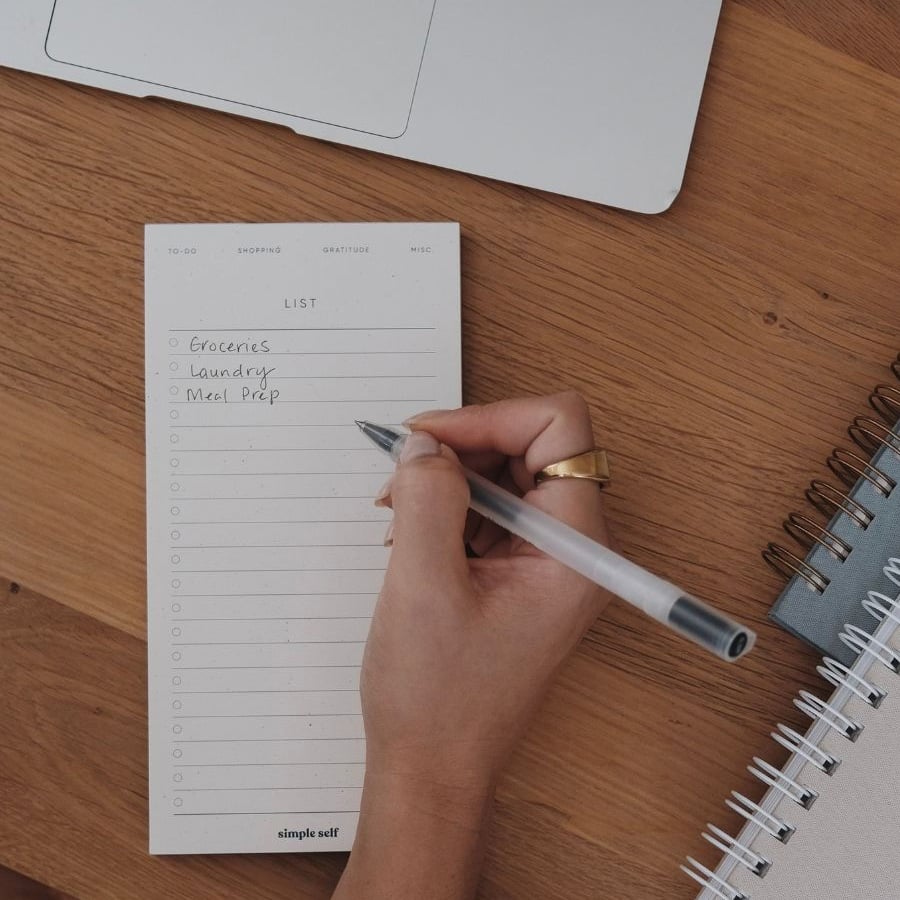 A hand writes a to-do list on a notepad with items: groceries, laundry, and meal prep. A pen, spiral notebook, and part of a laptop are visible on a wooden desk.