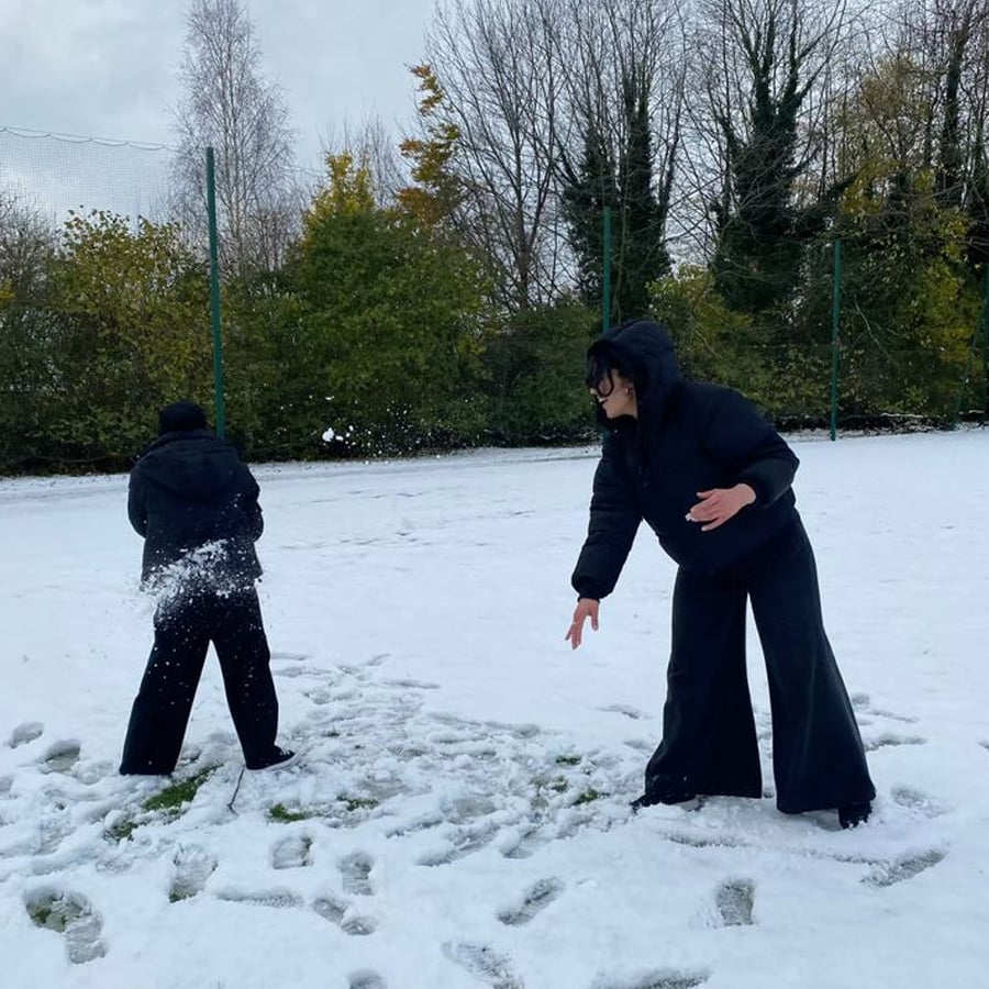 Two people dressed in dark winter clothes play in a snow-covered field. One throws a snowball while the other faces away, surrounded by snowy ground and trees in the background.