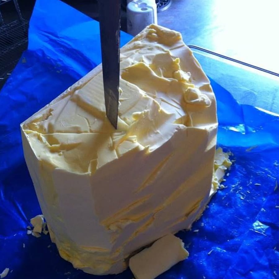 A large block of butter sits on a blue surface with a knife stuck vertically into its center. Bits of butter are shaved off around it.