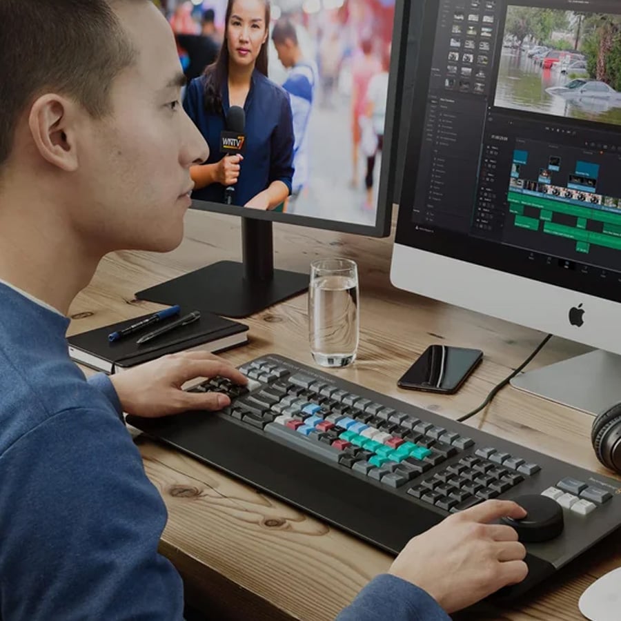 A person edits video footage on a desktop computer with a large keyboard, control knob, and two monitors; one shows a news reporter, the other displays a video editing program.