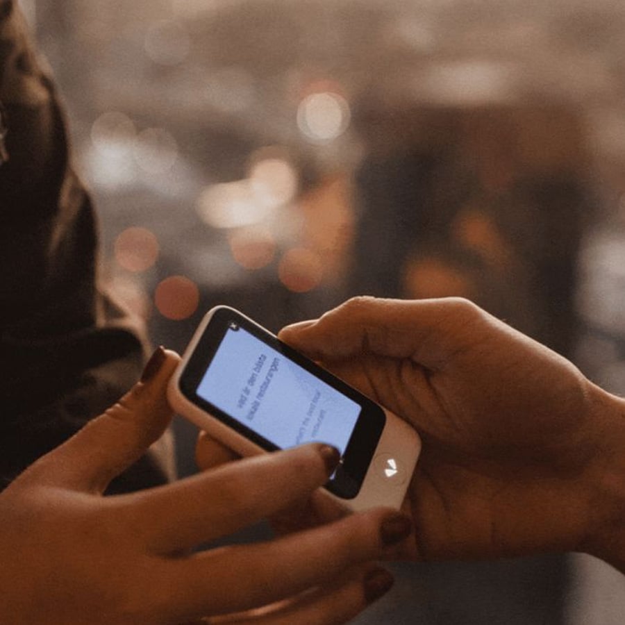 Two hands hold a small electronic device displaying text on its screen, with a blurred background of warm lights. The device appears to be used for communication or reading.