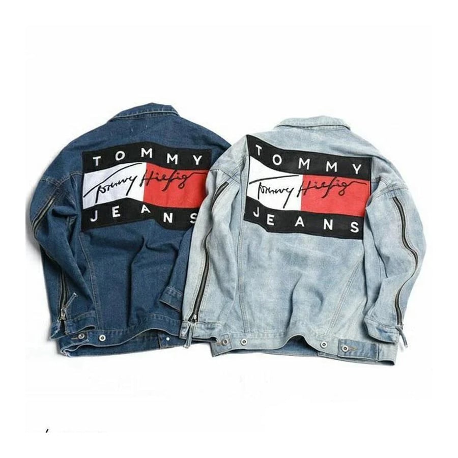 Two denim jackets, one dark blue and one light blue, both featuring a large “Tommy Jeans” logo patch with red, white, and black sections and cursive text on the back.