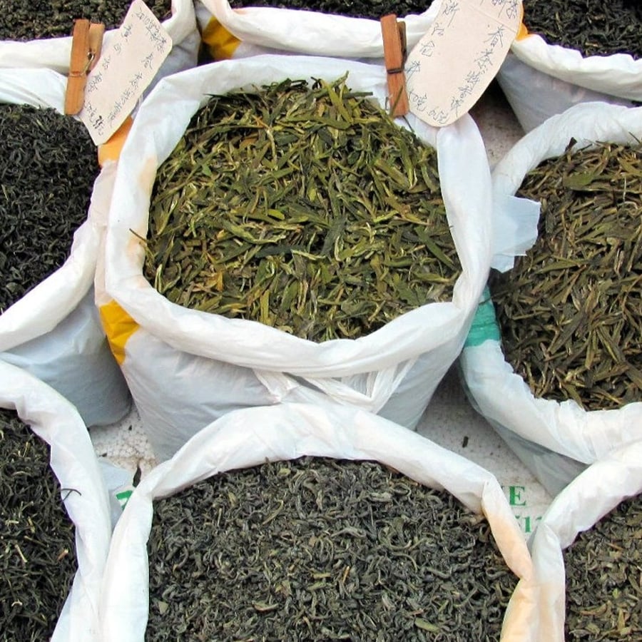 Large white sacks filled with different varieties of dried tea leaves are displayed at a market, with handwritten signs on paper attached to some bags.