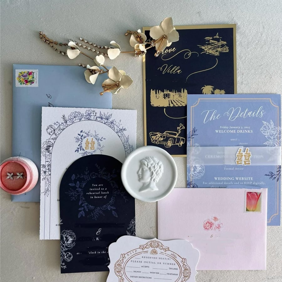 A flat lay of elegant wedding invitations, envelopes, a wax seal, a beaded bracelet, floral hairpins, and a pink ring box with two rings, all arranged on a light surface.