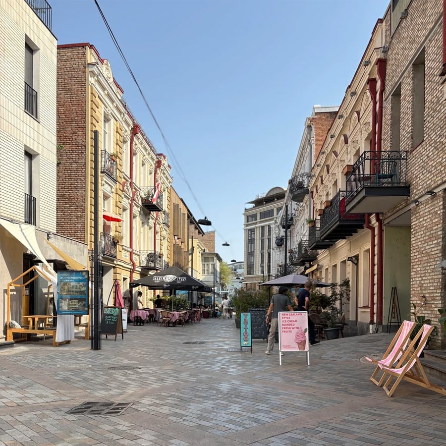 A cobblestone pedestrian street with outdoor cafes, pink chairs, and signs. People are sitting and walking. The street is lined with historic buildings under a clear blue sky.