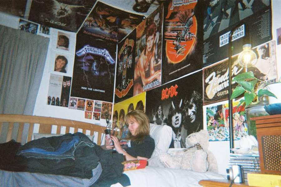 A person lies on a bed in a room covered with rock band posters. Clothes and a stuffed animal are on the bed, and a lamp and plant are on a nearby nightstand. The person looks at a phone.