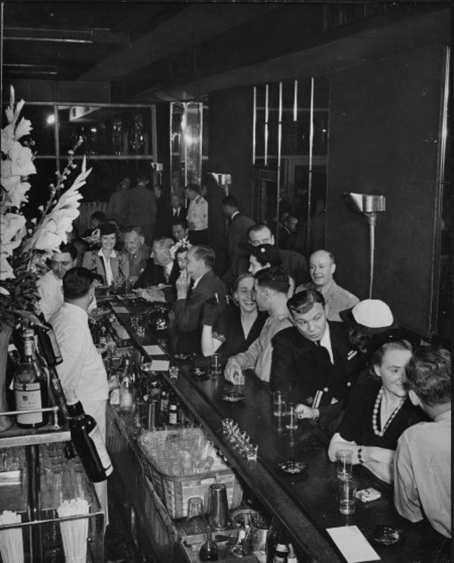 A crowded, lively bar scene from the mid-20th century shows people socializing and bartenders serving drinks. Patrons in suits and dresses are engaged in conversation, with flowers and bottles visible on the counter.