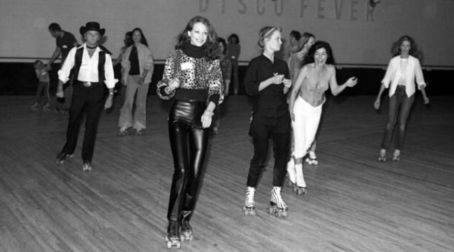 People roller skating indoors on a wooden floor, smiling and having fun. The setting has a retro vibe, with some wearing disco-style outfits. The words “DISCO FEVER” are visible on the wall in the background.