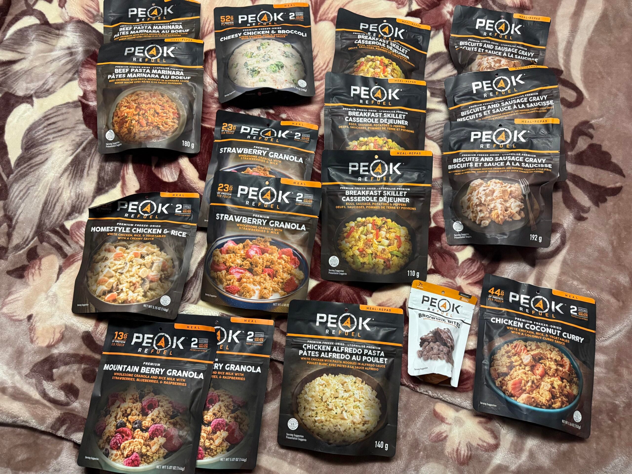 Fourteen Peak 2 meal pouches and one Peak snack pack are arranged on a patterned blanket, displaying various freeze-dried meal options like chicken curry, granola, and pasta.