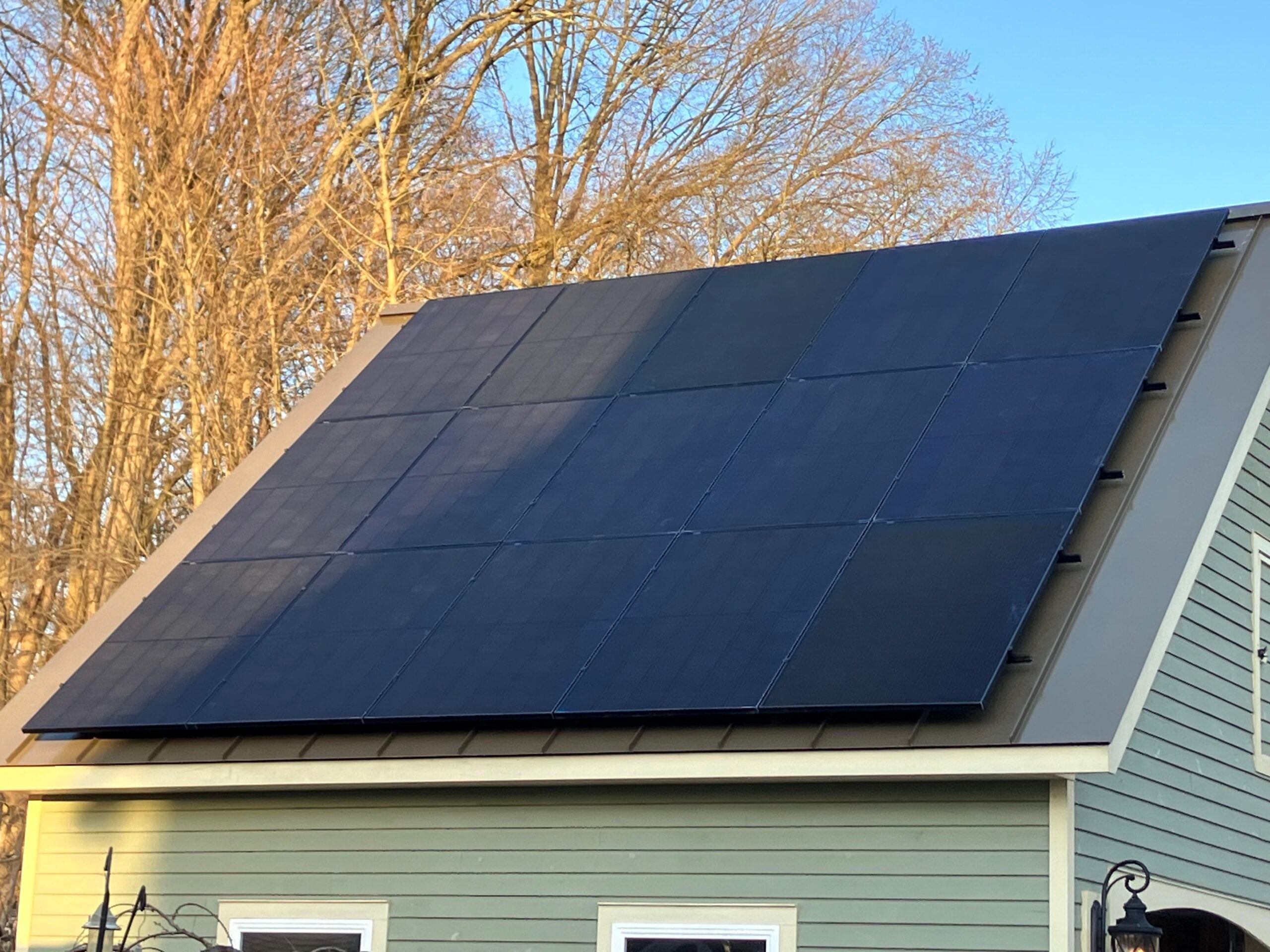 Rooftop with large black solar panels installed on a sloped green house, with bare trees and a clear blue sky in the background.