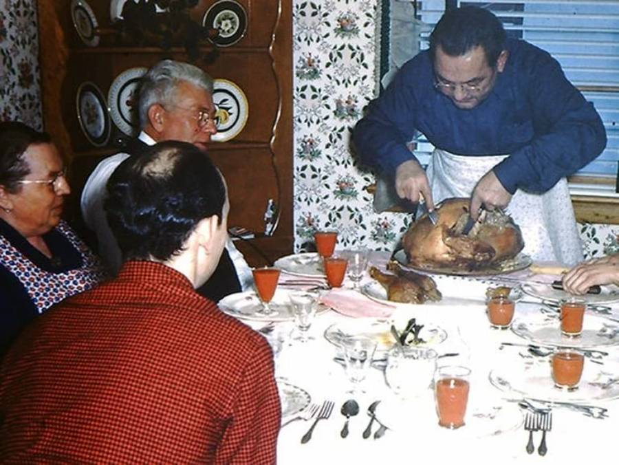 A man carves a turkey at a dining table surrounded by three seated people. The table is set with glasses of red drink and plates. The room has floral wallpaper and a cabinet with plates.