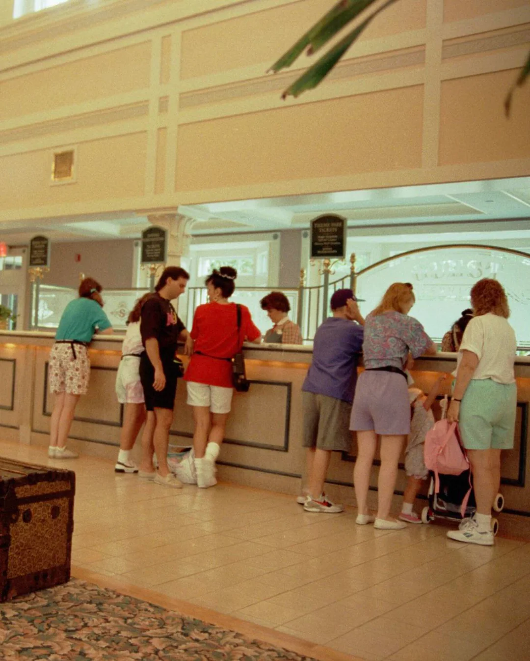 Several people dressed in colorful 1990s clothing stand at a hotel reception counter, checking in or speaking with staff. The lobby has bright lighting and a patterned tile floor.