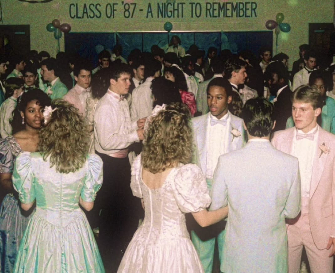High school students in pastel formal wear dance at a prom under a banner reading “Class of ’87 - A Night to Remember.” Balloons hang on the wall and many couples are visible.