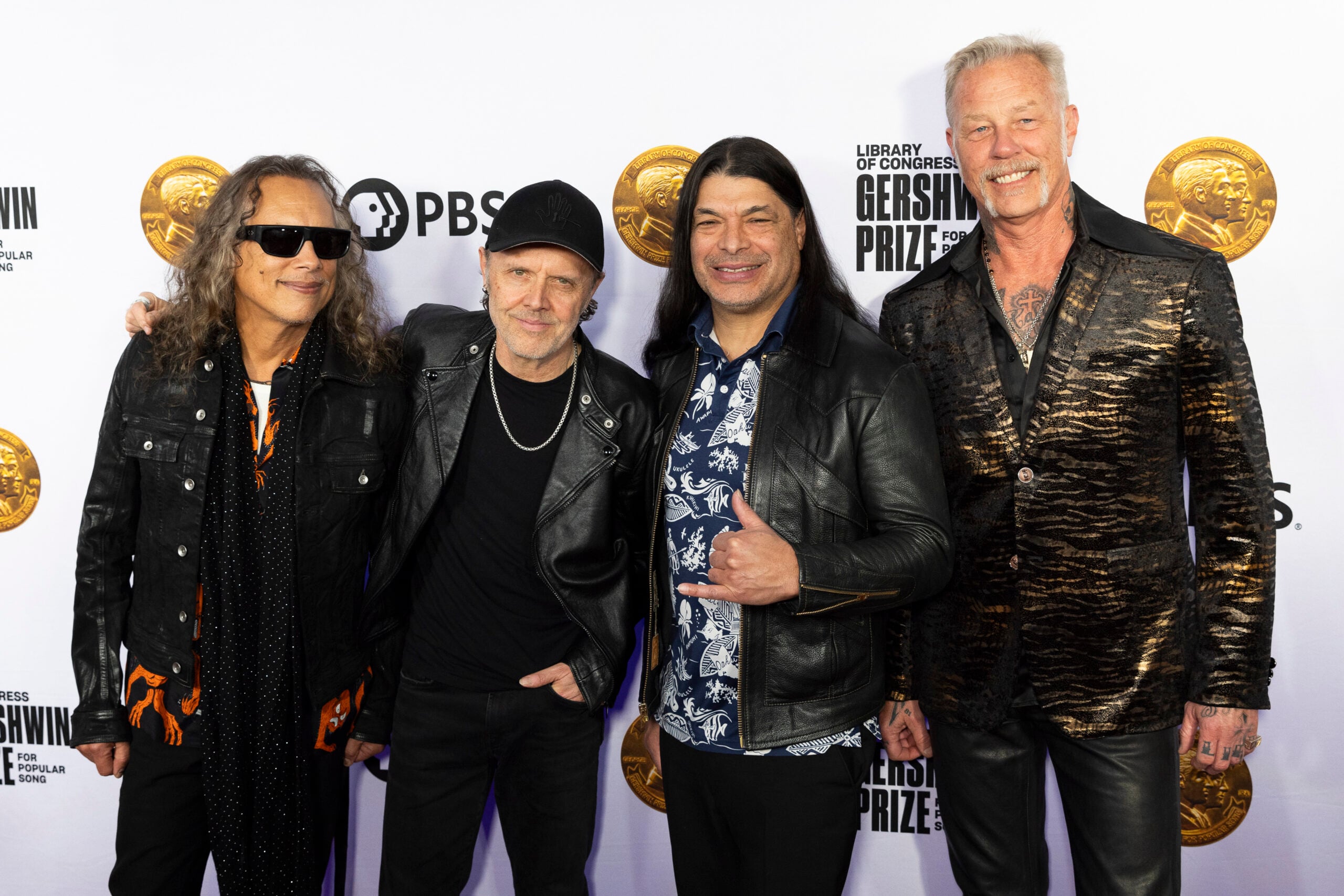 Four men stand together, smiling, on a red carpet in front of a backdrop with gold medallions and text. They are wearing black jackets, and one wears sunglasses.