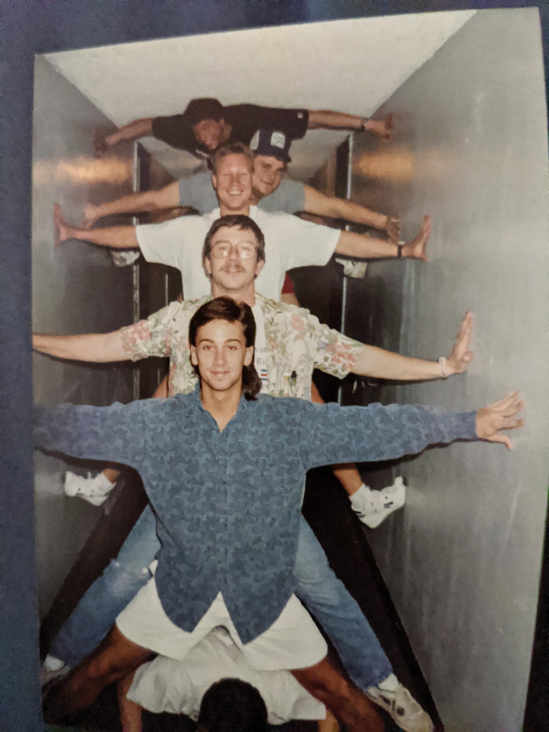 Five people are stacked vertically in a narrow hallway, each with arms and legs outstretched, pressing against opposite walls, creating a human tower. All are smiling and casually dressed.