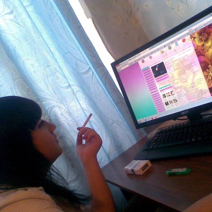 A person holding a cigarette sits at a desk, looking at a colorful website on a computer monitor. A pack of cigarettes and a lighter are on the desk. Light blue curtains are in the background.