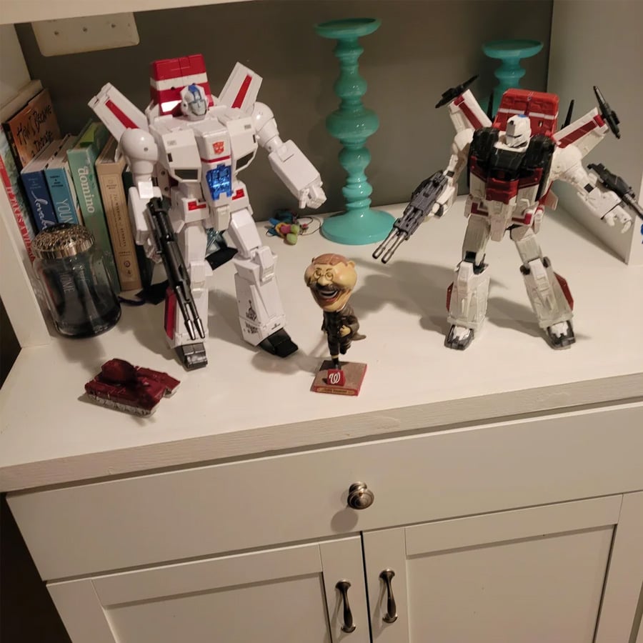 Two Transformers action figures, a bobblehead, two turquoise candle holders, books, a glass container, and a small tray with items are displayed on a white shelf with a cabinet below.