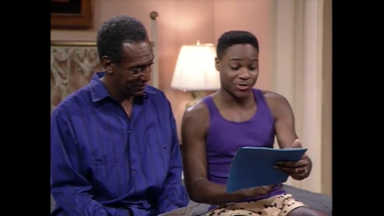 Two men sit close together indoors; one wears a blue shirt and looks at the other, who is in a purple tank top and holding blue papers, reading aloud. A lit lamp is in the background.