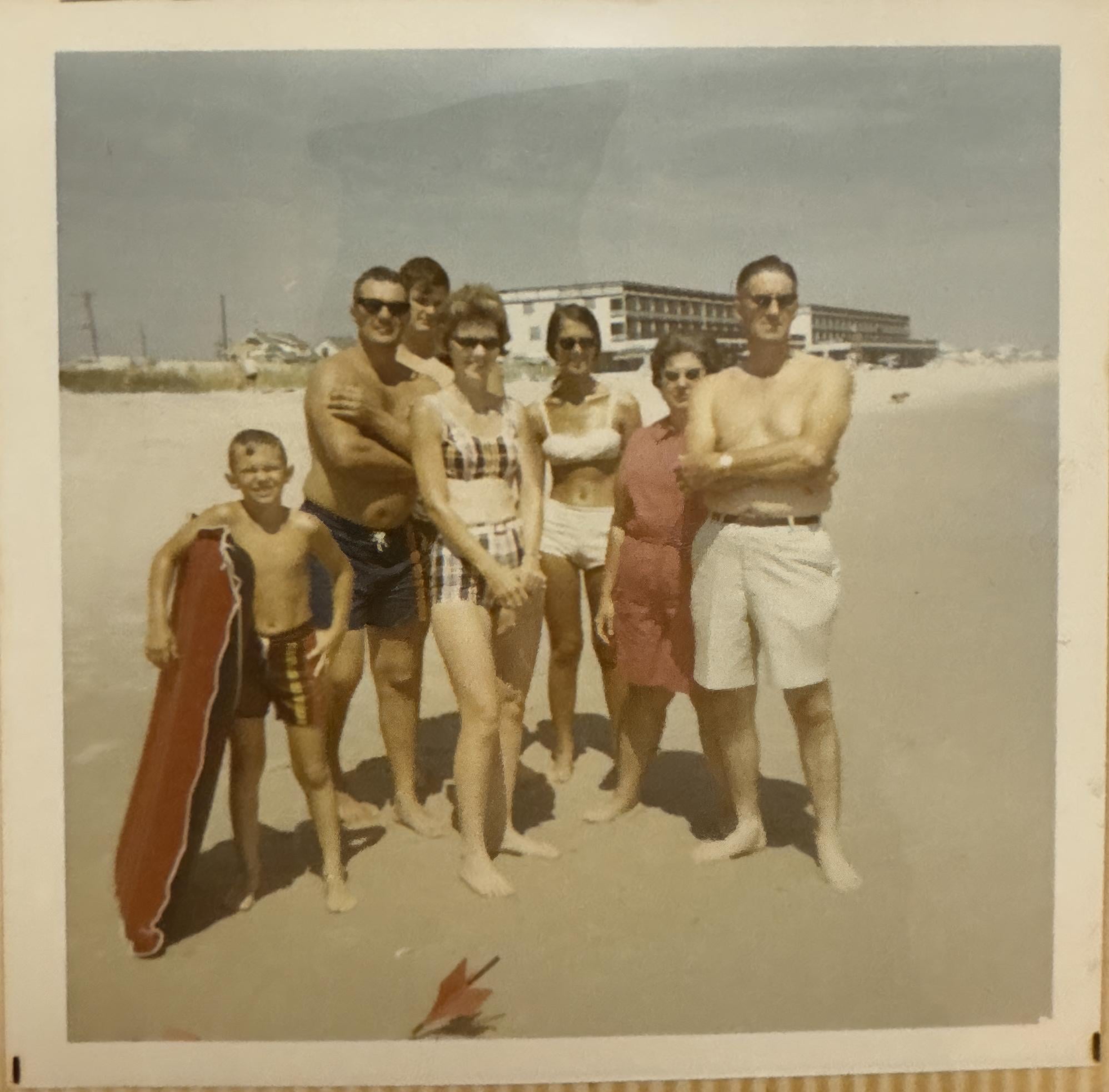 Six people in vintage swimwear stand on a sandy beach, posing for a photo. A young boy holds a red float. There’s a large building and blue sky in the background, suggesting a summer day at the seaside.