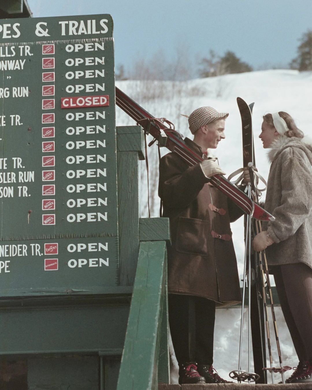 Two people in winter clothing stand by a ski slopes and trails sign, holding skis and poles, with a snowy hill in the background. Most trails on the sign are marked as open except one that is closed.