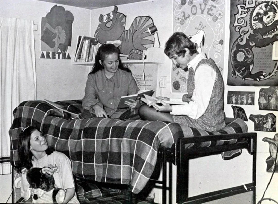 Three young women are in a decorated dorm room; two sit on a top bunk bed reading, while one sits below on the floor holding a stuffed animal. Posters and artwork are visible on the walls around them.