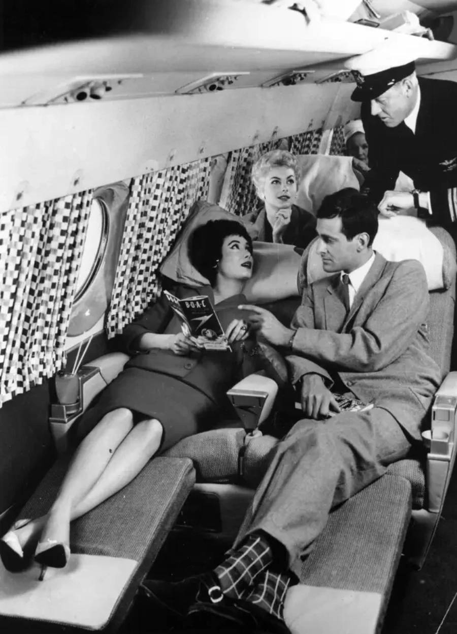 A black-and-white photo of four people in an airplane cabin. A woman reclines with a magazine, a man sits beside her, another woman sits behind, and a pilot leans in to talk to them.