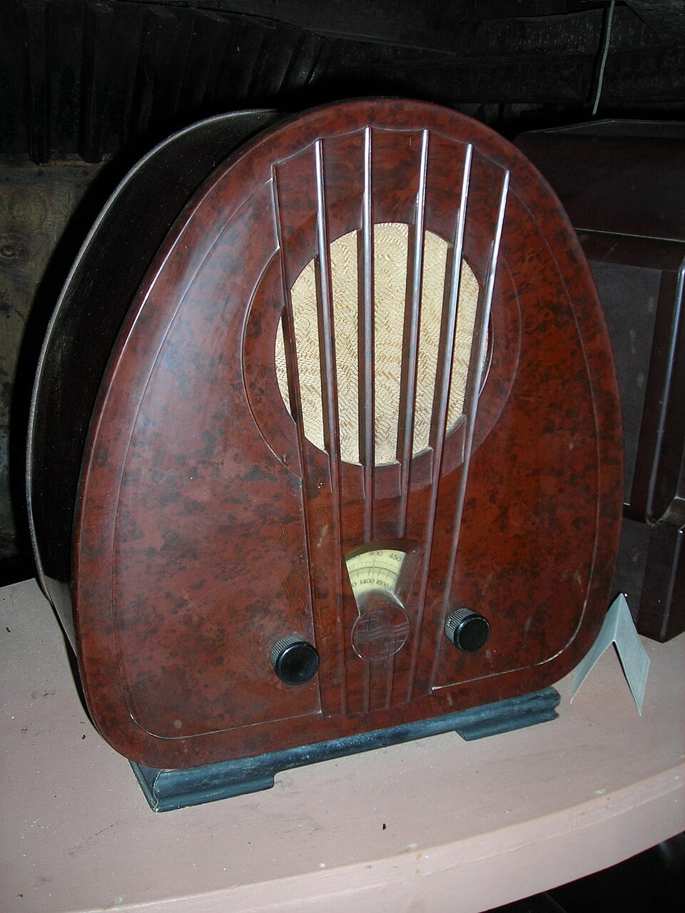 A vintage brown tabletop radio with a rounded top, vertical grill lines, a circular speaker, two knobs, and a small display, sitting on a light-colored surface.