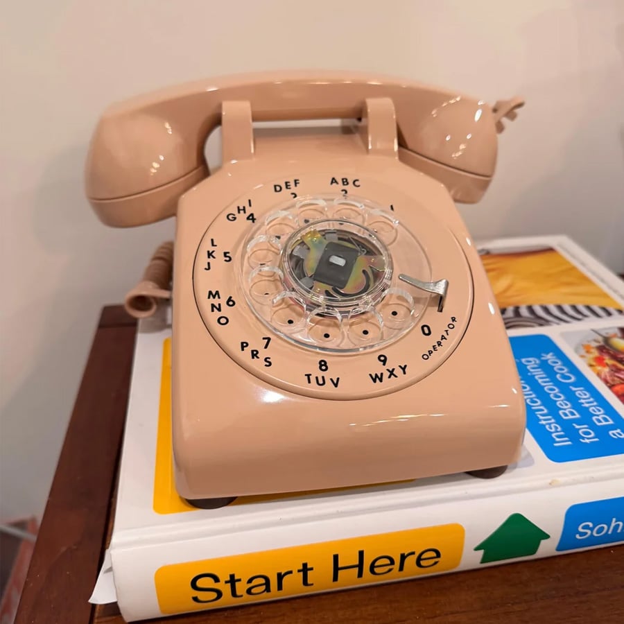 A beige rotary dial telephone sits on a book titled “Start Here” with a cookbook partially visible beneath it on a wooden surface.