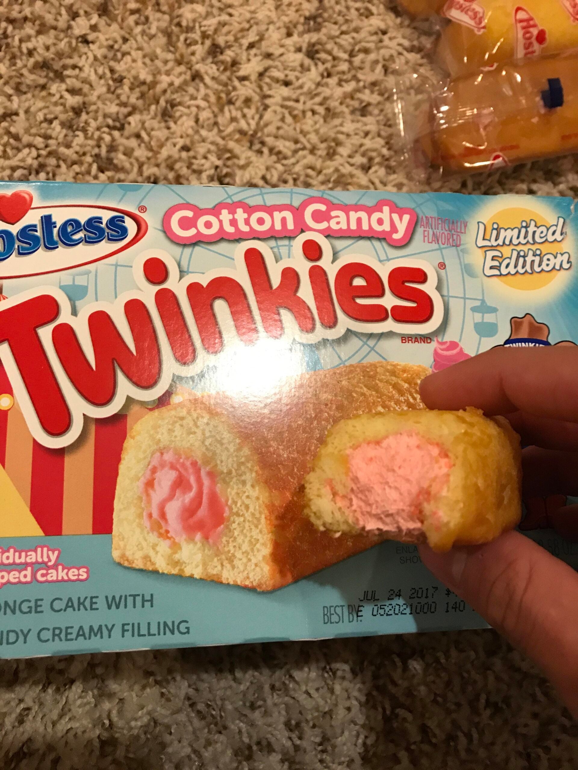 A hand holds a half-eaten cotton candy flavored Twinkie with pink filling in front of its colorful box on a carpeted floor.