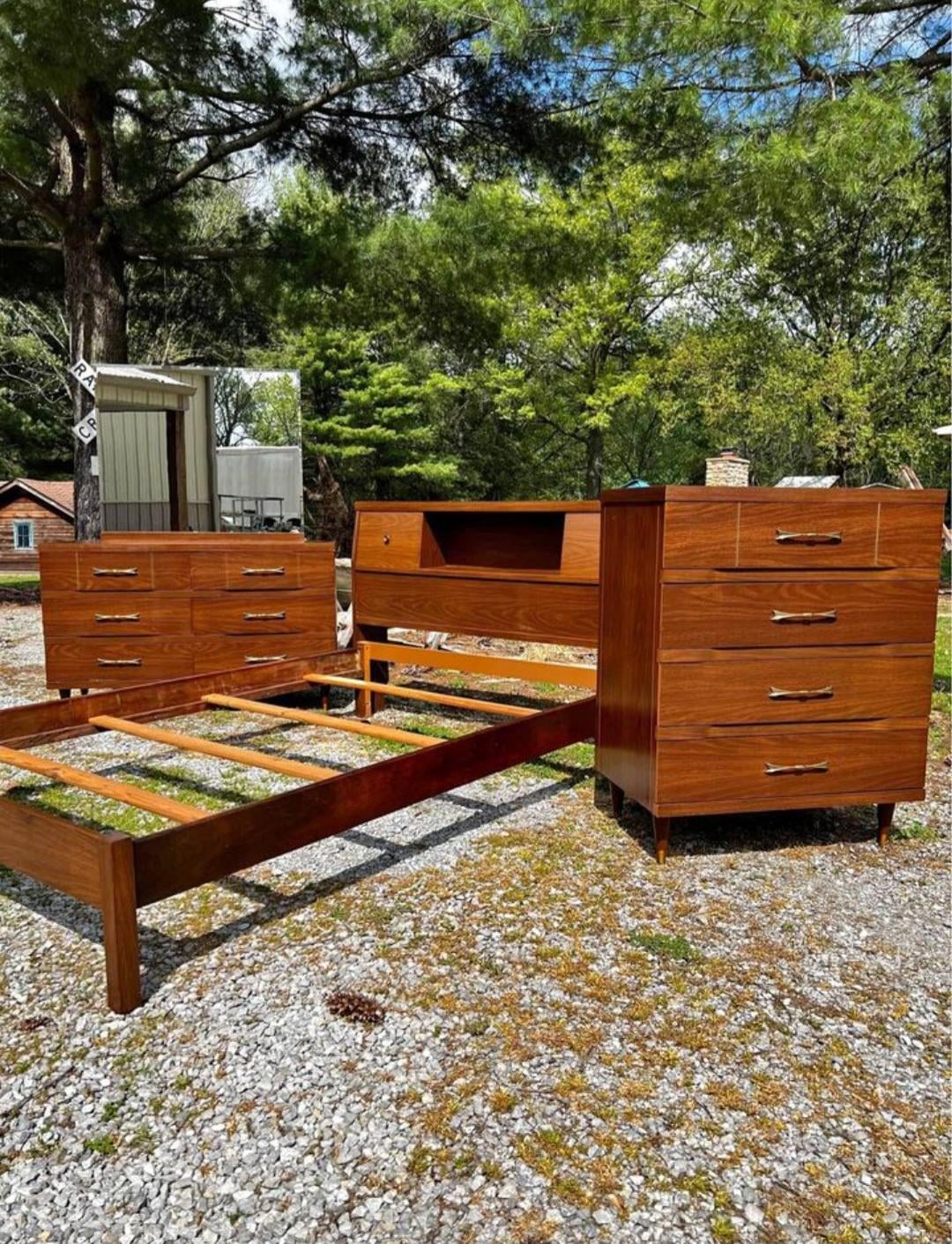 Four matching mid-century modern wooden dressers and a bed frame are arranged outdoors on a gravel surface, with trees and green grass in the background.