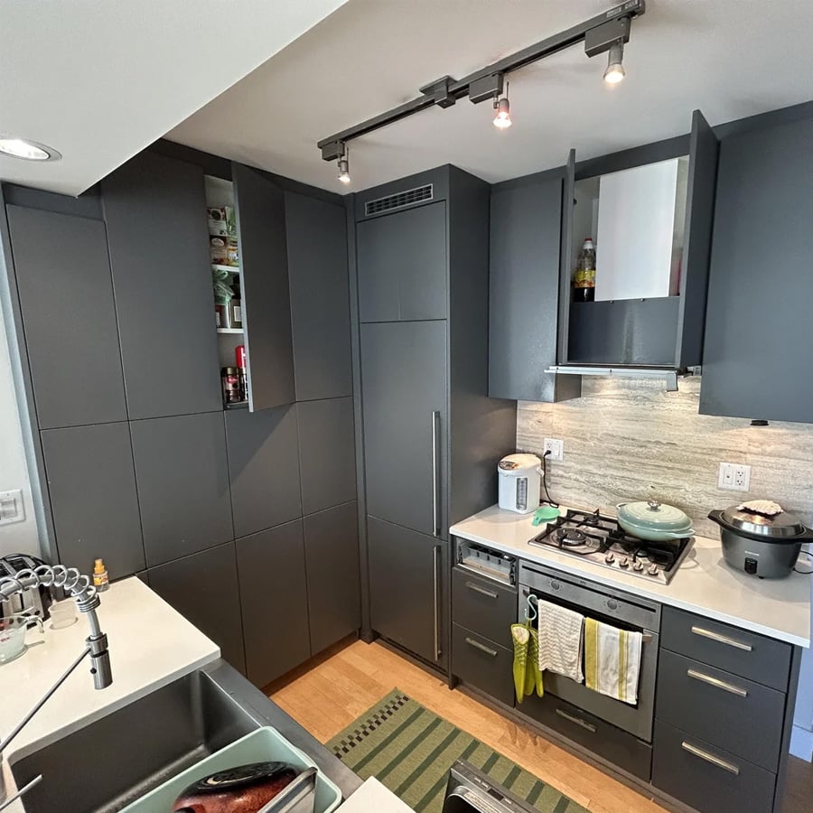 Modern kitchen with dark gray cabinets, some open to reveal pantry items, appliances on countertops, green-striped rug, gas stove with pots, and dish rack by the sink. Track lighting is mounted on the ceiling.