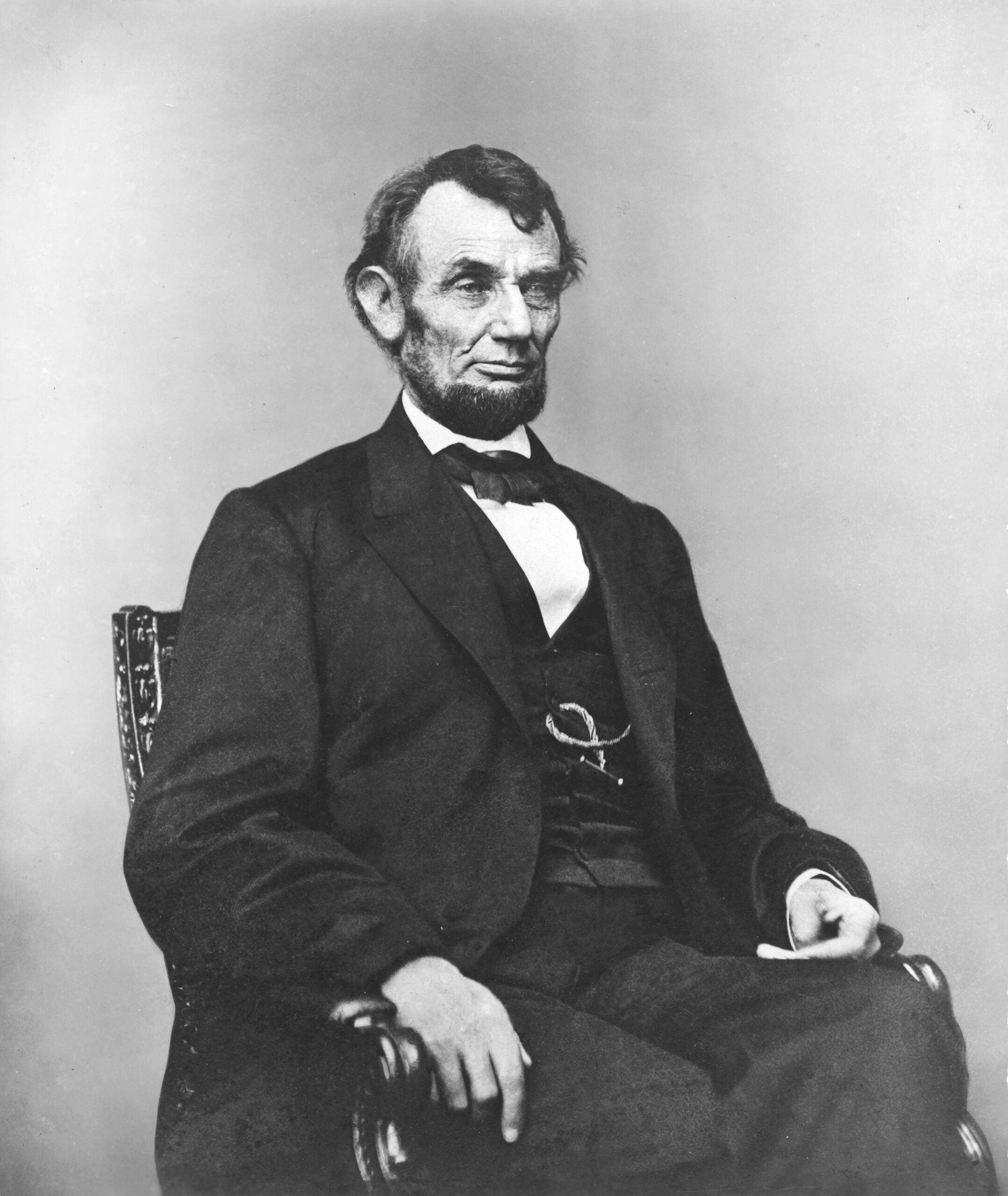 A black-and-white portrait of a man with a beard, wearing a suit and bow tie, seated on a carved chair, looking slightly to the side against a plain background.