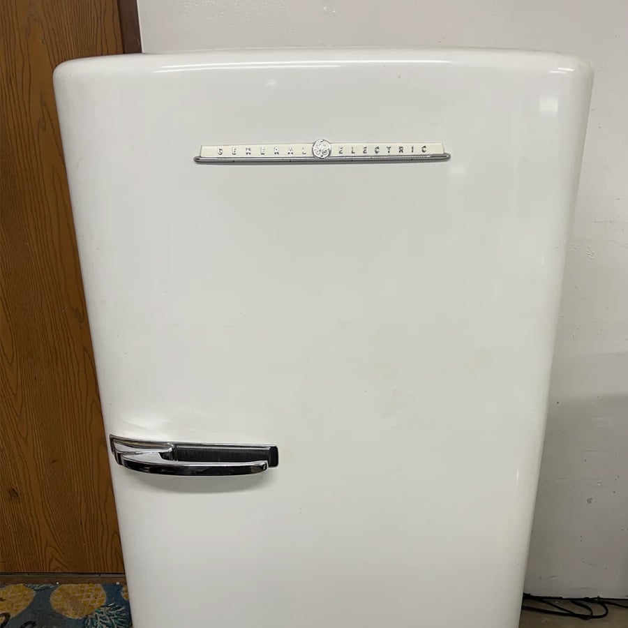 A vintage white General Electric refrigerator with a chrome handle and label, standing against a white wall and wooden door. Only the upper part of the appliance is visible.