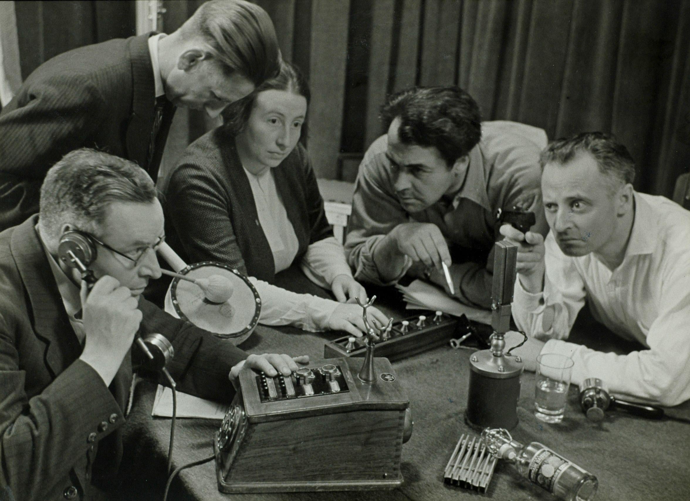 Five people gather around a table with old-fashioned radio equipment, microphones, and papers, appearing focused and engaged in discussion or operation, possibly working on a broadcast or scientific experiment.