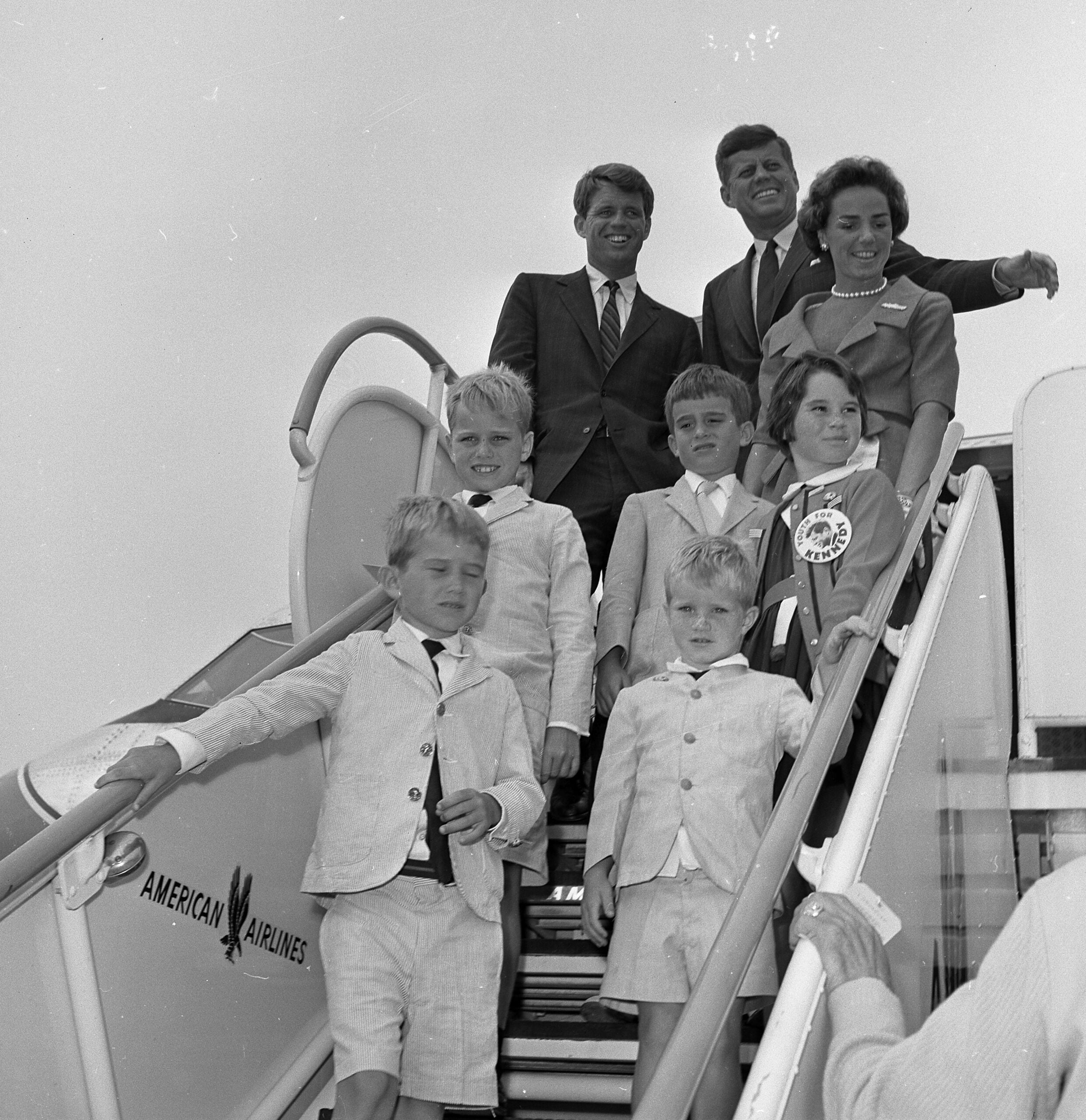 A group of adults and children, formally dressed, pose and smile while standing on the stairs of an American Airlines airplane.