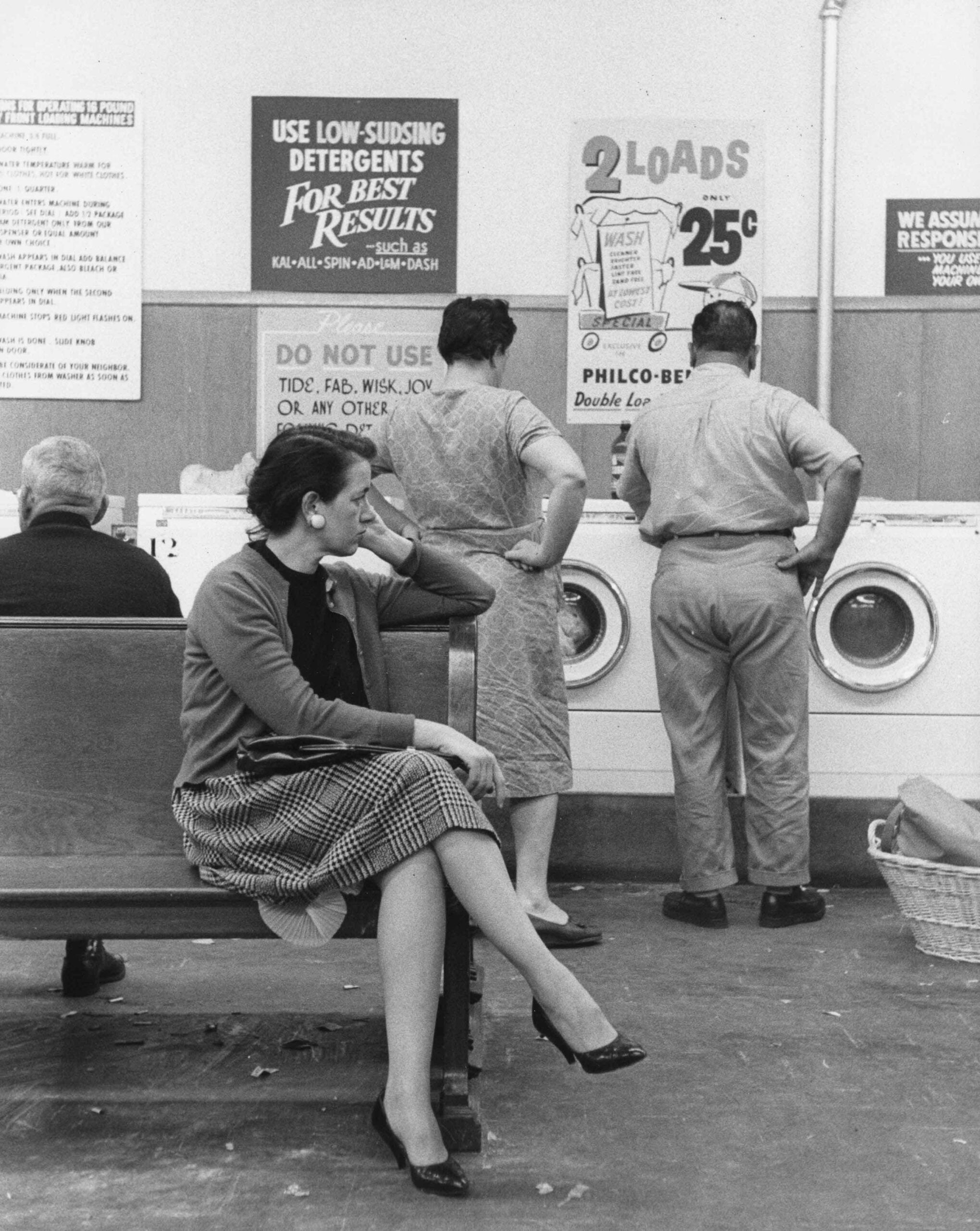 Black-and-white photo of people at a laundromat. A woman sits on a bench in the foreground, while others stand by washing machines. Laundry-related signs are posted on the wall.