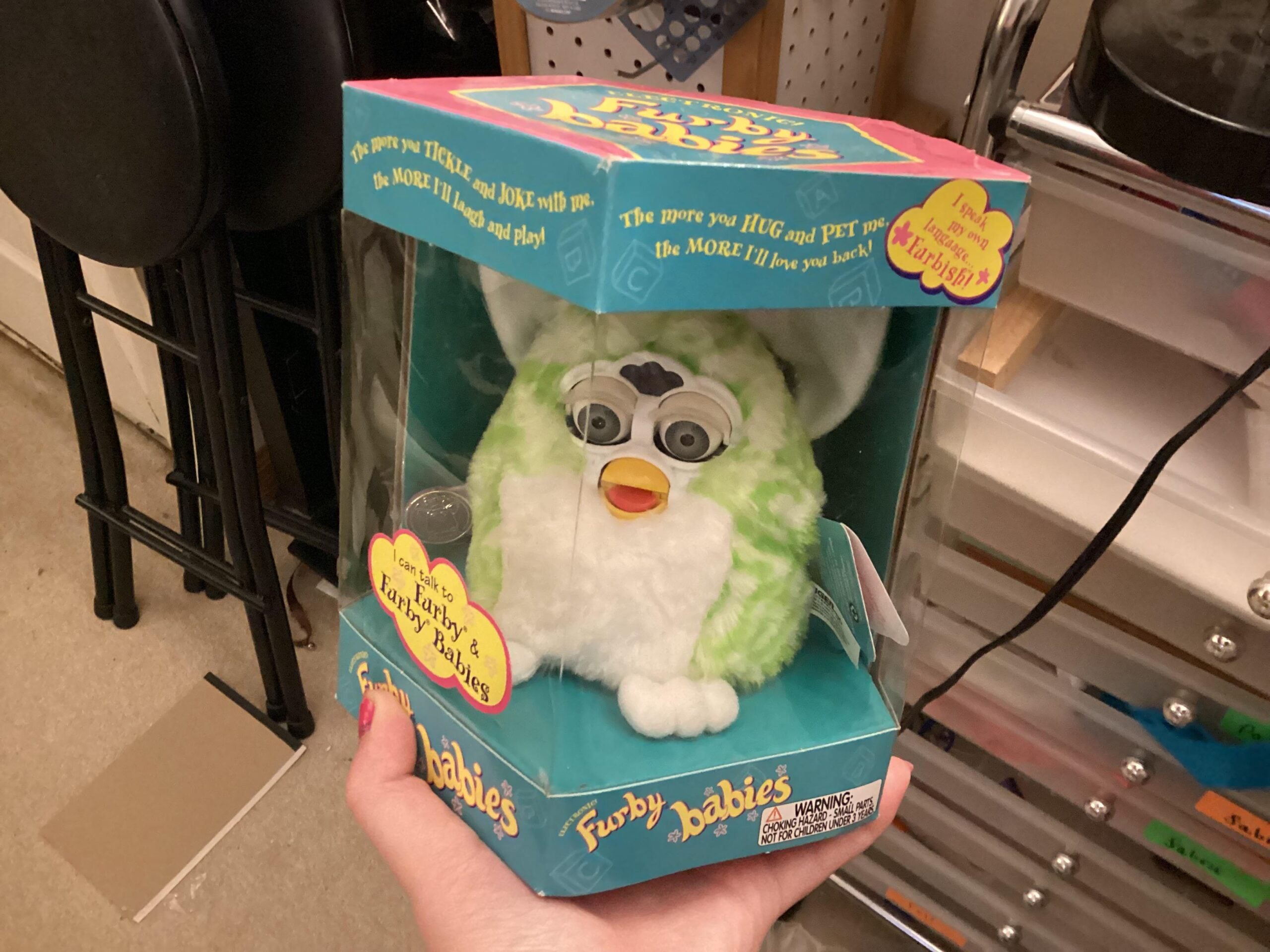 A hand holds a boxed green and white Furby Babies toy in packaging with colorful text, displayed in a room with shelves and other items in the background.