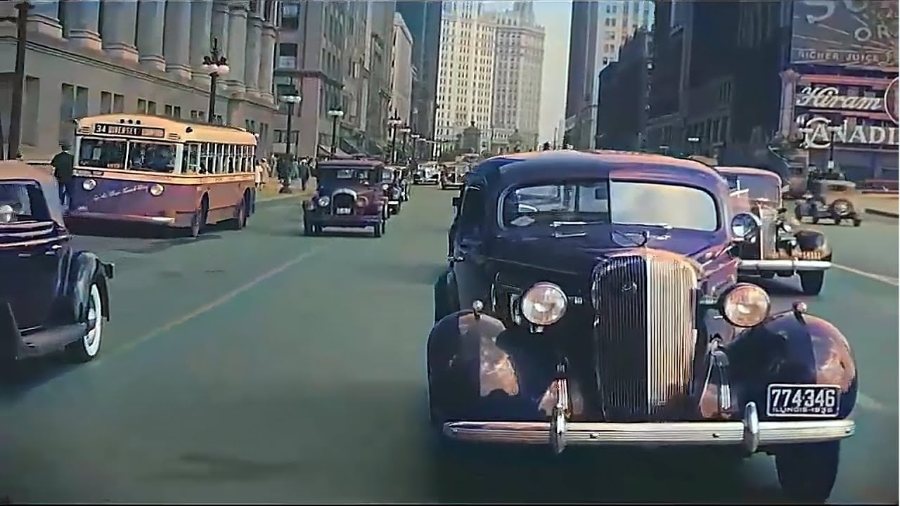 Vintage cars and an old-fashioned bus drive down a bustling city street lined with tall buildings, evoking a mid-20th century urban scene.