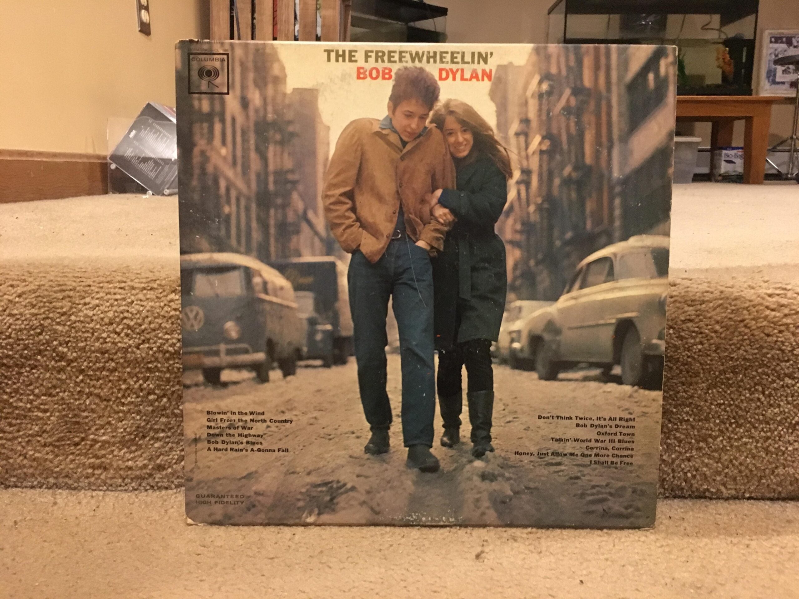 Bob Dylan’s album “The Freewheelin’ Bob Dylan” cover shows him walking arm-in-arm with a woman down a city street lined with parked cars and old buildings.