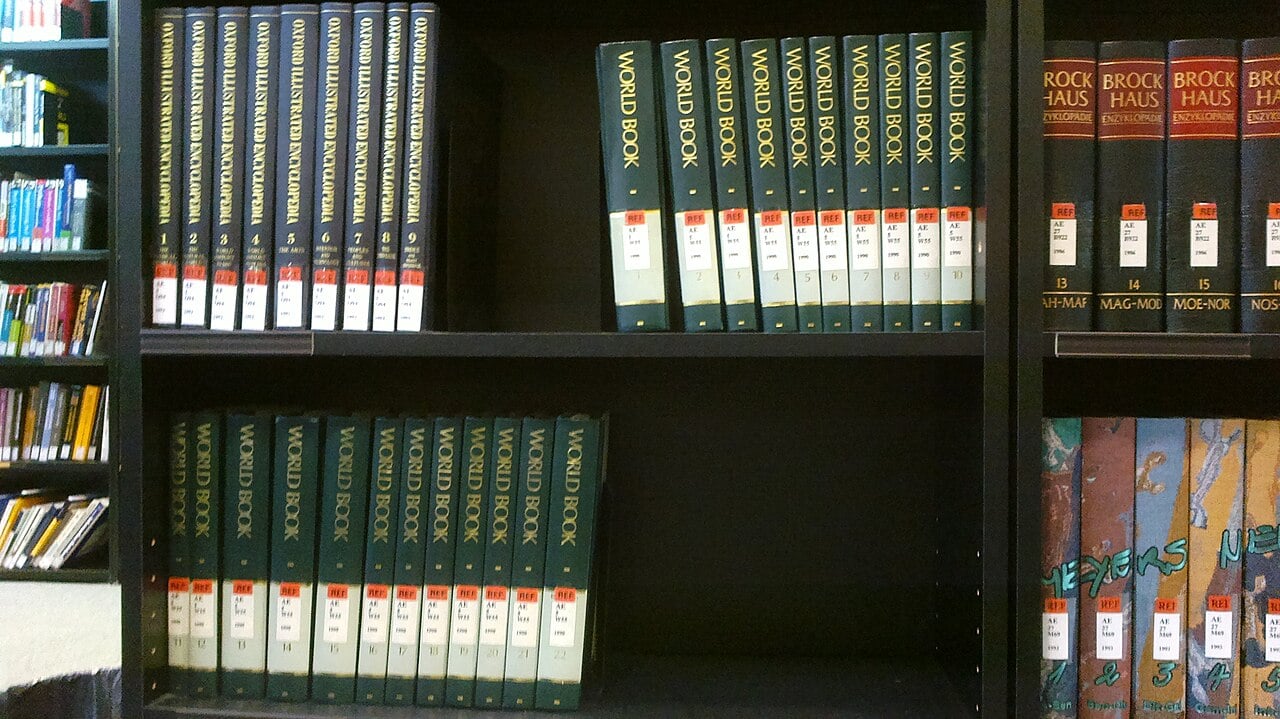Shelves in a library display multiple volumes of the World Book Encyclopedia and other reference books, arranged neatly in alphabetical and numerical order.