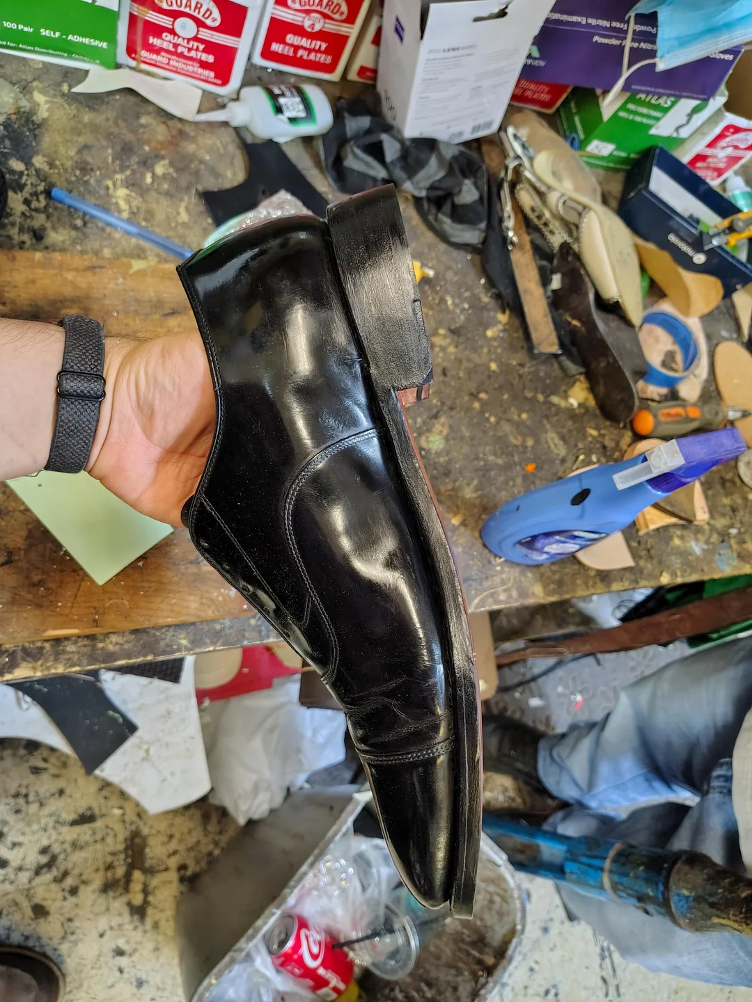 A person holds a black, shiny dress shoe in a workshop with tools, shoe materials, and cluttered work surfaces visible in the background.